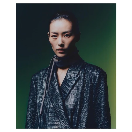 Portrait of an Asian woman in a reptile-textured black blazer, with a dark gray scarf, against a gradient green background.