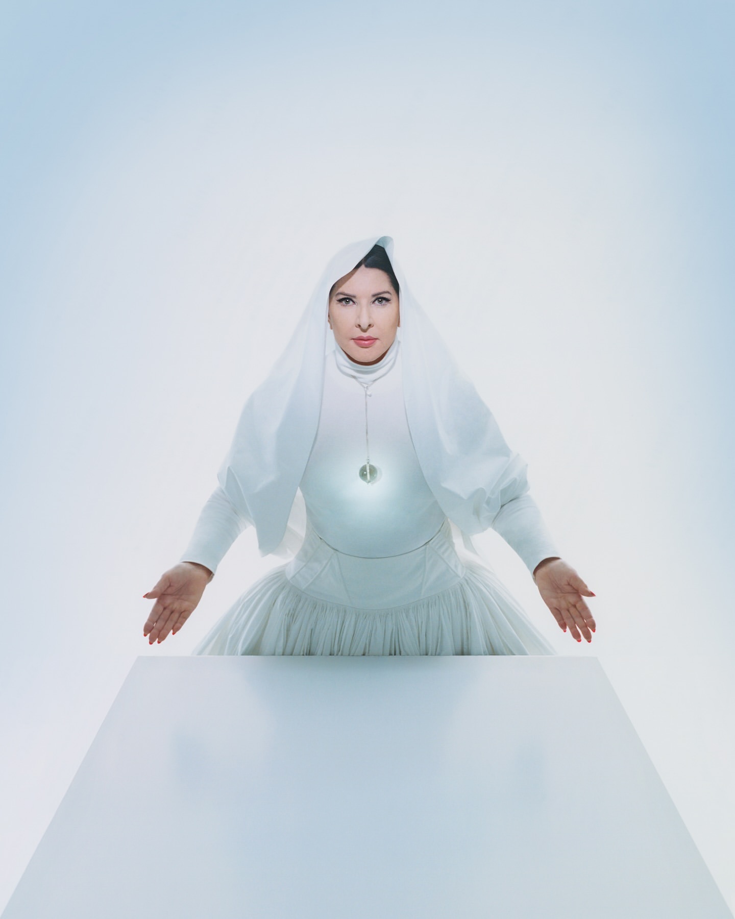 Portrait of adult female in white dress and head covering standing behind a white table against a bright white and pale blue background.
