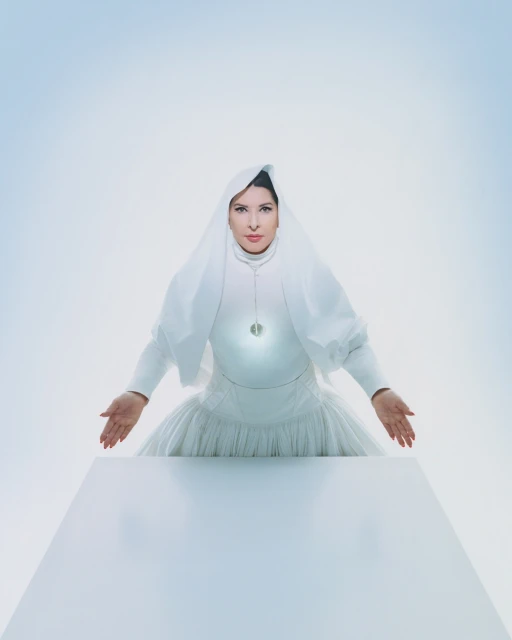 Portrait of adult female in white dress and head covering standing behind a white table against a bright white and pale blue background.