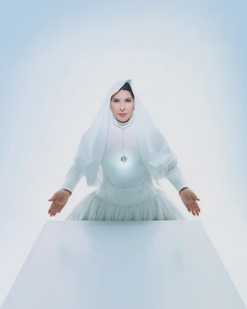 Portrait of adult female in white dress and head covering standing behind a white table against a bright white and pale blue background.