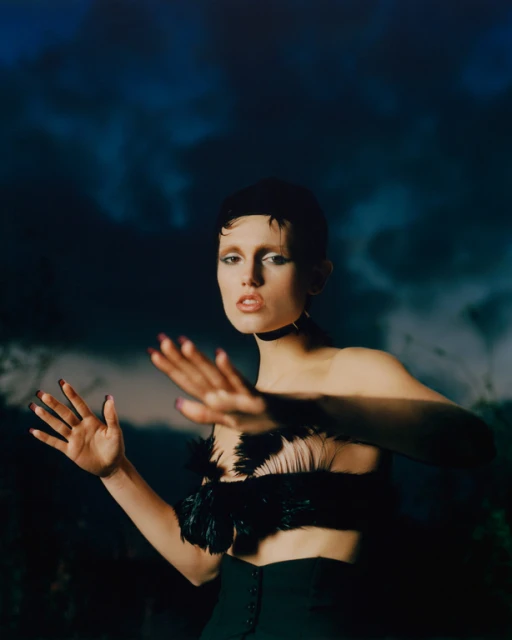 Portrait of a young adult female model in feather top with expressive hand gestures against a blurred blue and green backdrop.
