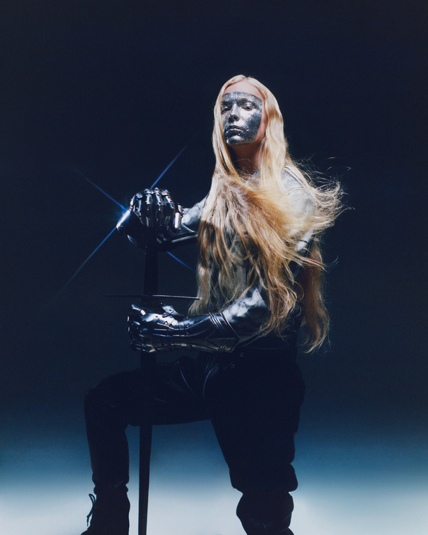Silver-skinned person with long blonde hair poses with a sword against a shadowy backdrop, creating a futuristic warrior aesthetic.