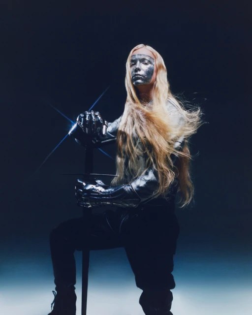 Silver-skinned person with long blonde hair poses with a sword against a shadowy backdrop, creating a futuristic warrior aesthetic.
