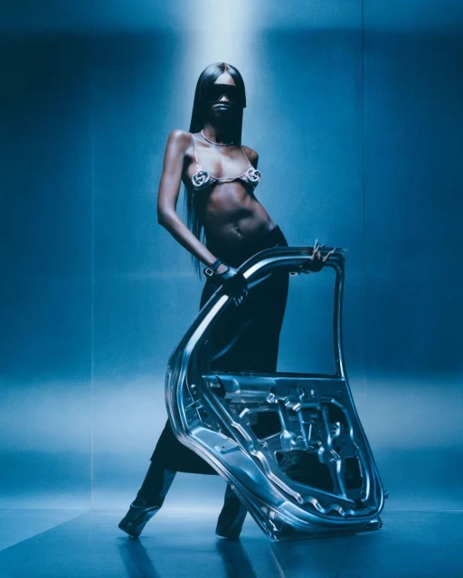 A woman with long dark hair poses in a bikini top and black pants, holding a silver car door against a gradient blue background.