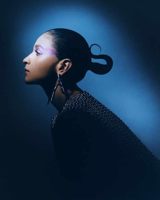 Portrait of a stylish young woman with creative eye makeup, wearing a studded top and statement earrings, against a cyan background.
