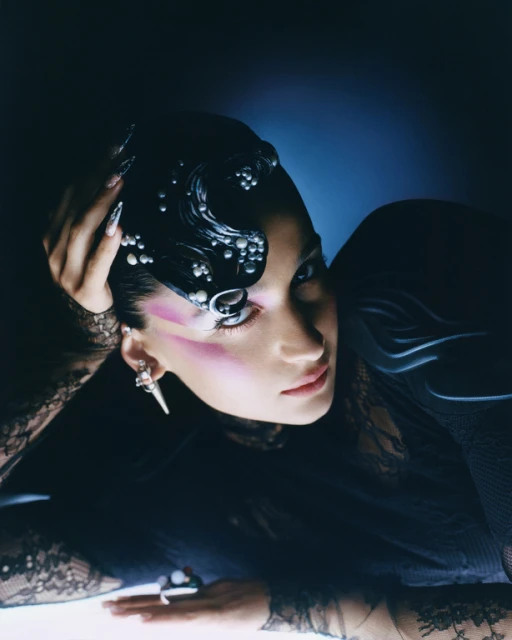 Elegant woman with creative makeup, adorned with pearl-like beads and wearing sheer black lace, poses in moody, low-key lighting.