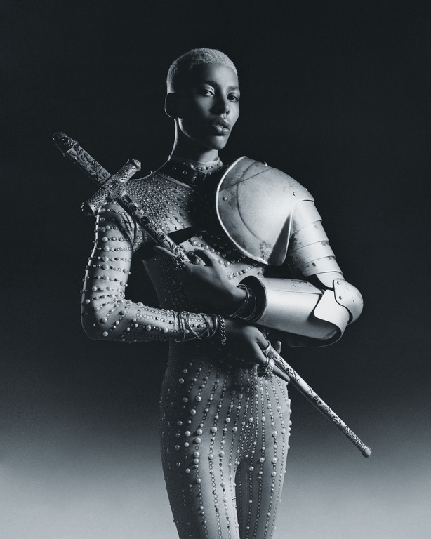 Portrait of a person with short hair wearing a studded body suit with partial armor and holding a sword in a dark studio setting.