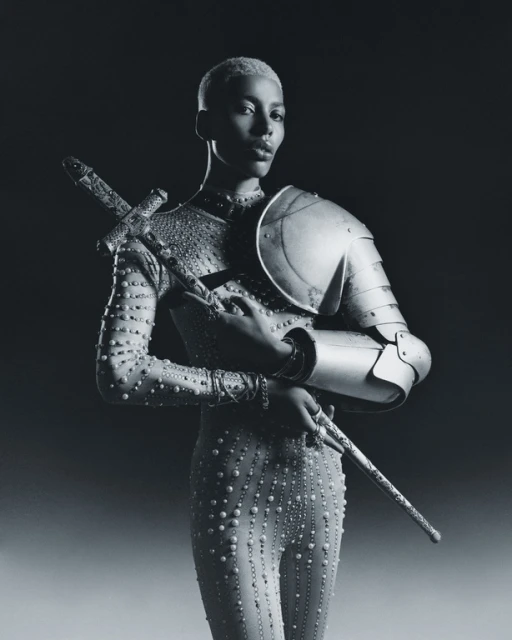 Portrait of a person with short hair wearing a studded body suit with partial armor and holding a sword in a dark studio setting.