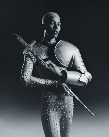 Portrait of a person with short hair wearing a studded body suit with partial armor and holding a sword in a dark studio setting.