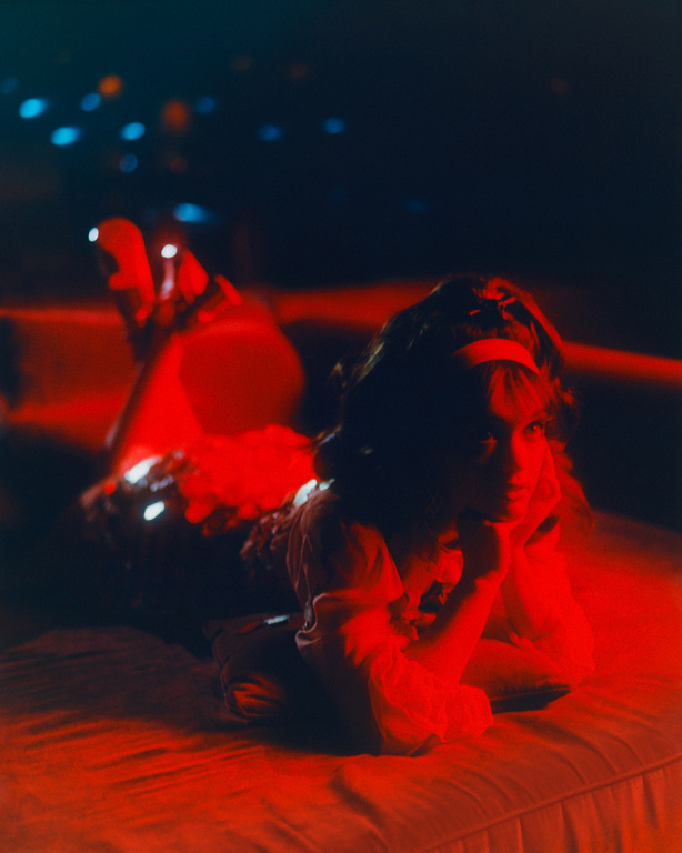 Child lying on a bed with a red light, looking up. Dark setting with blue lights in background. Bedding, hairband, and skin tones are red.
