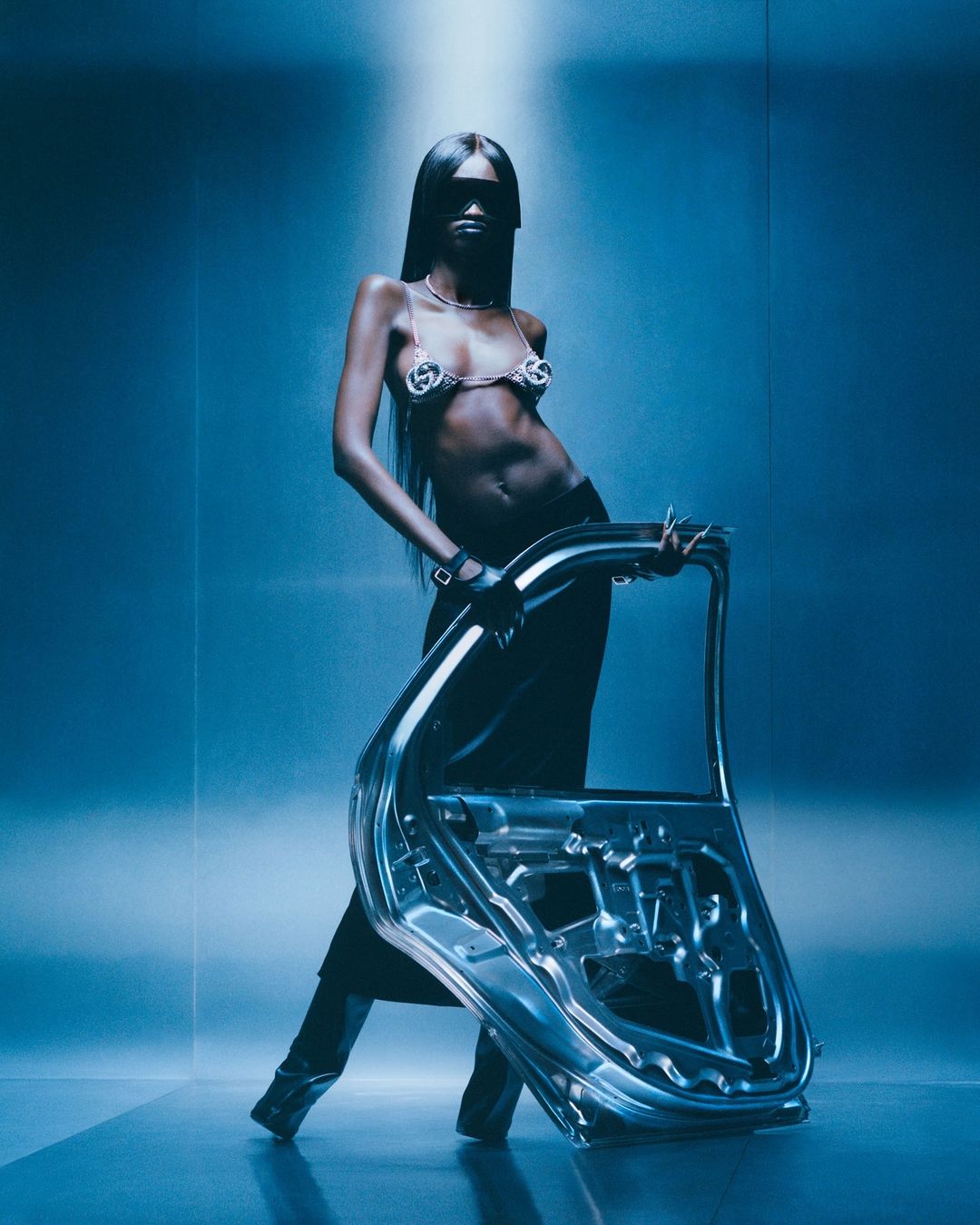 A woman with long hair and a metal bra poses with a car door in a studio with blue lighting, showcasing fashion and automotive elements.