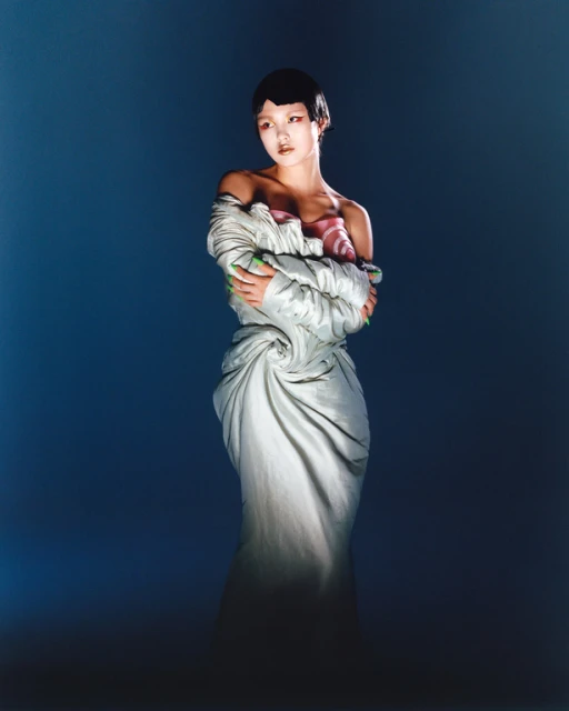 Elegant woman with short dark hair wears a light-grey couture gown in a studio setting against a diffused blue background, in a fashion shot.