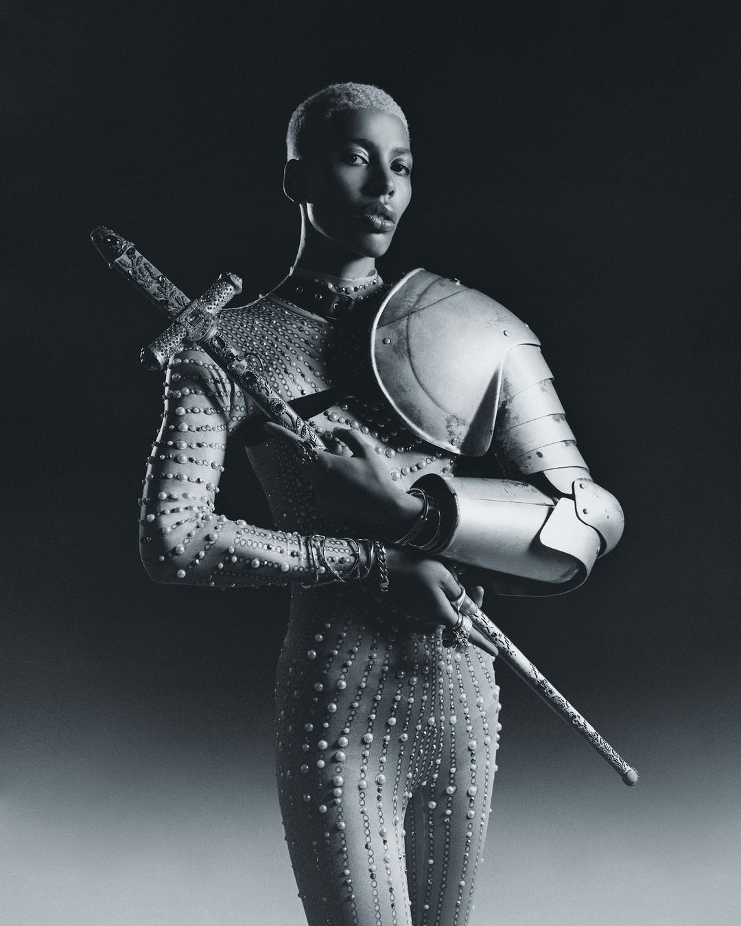 An adult female model with short blond hair wears a studded bodysuit and metal armour while holding a sword in a studio shot.