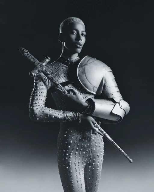 An adult female model with short blond hair wears a studded bodysuit and metal armour while holding a sword in a studio shot.
