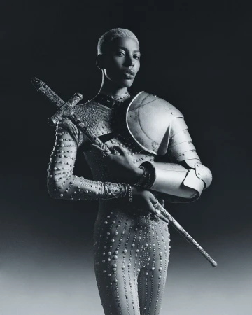 An adult female model with short blond hair wears a studded bodysuit and metal armour while holding a sword in a studio shot.