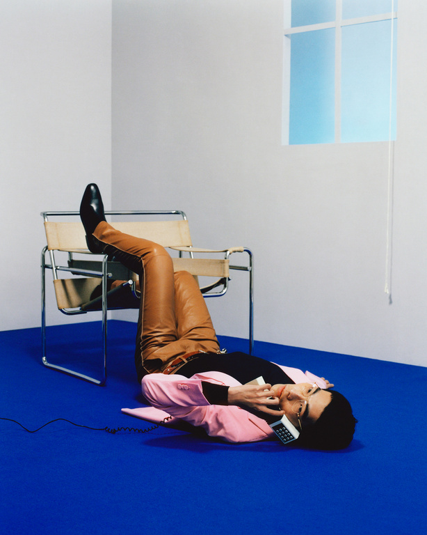 Stylish adult male lying on blue floor in pink suit and brown leather pants talking on a vintage phone next to a chair and window.