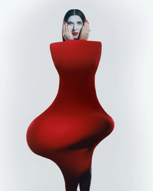 Model poses in a striking red sculptural dress, hands framing her face against a stark white background, creating a high-fashion editorial image.