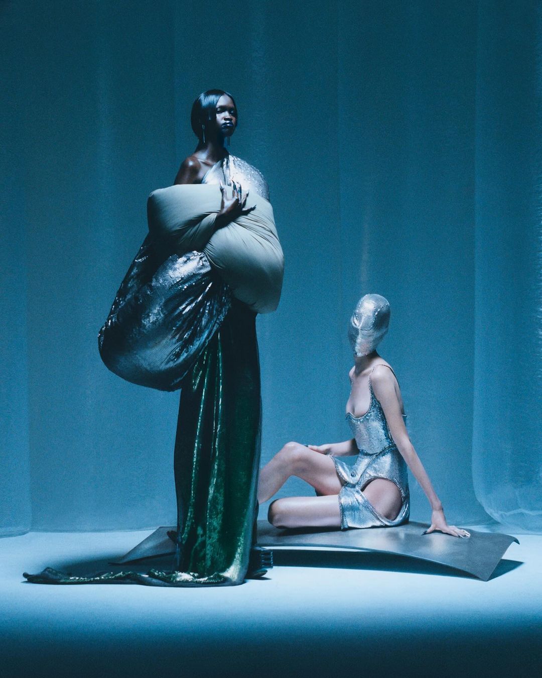 Two fashion models in a photo studio wearing avant-garde metallic dresses in a cool-toned, blue-lit studio. One model is seated, the other is standing.