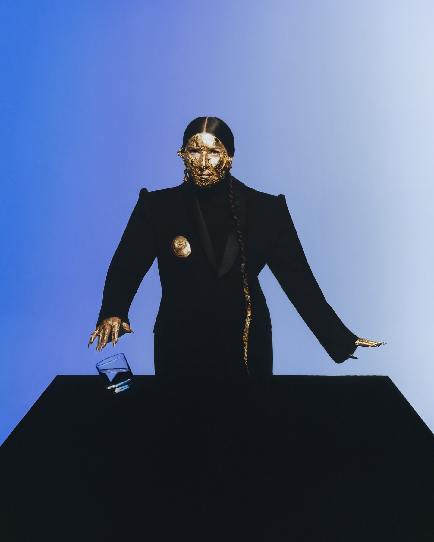 Model with a gold face mask in a black suit stands behind a black table. The model is reaching for a glass with her gold hand.