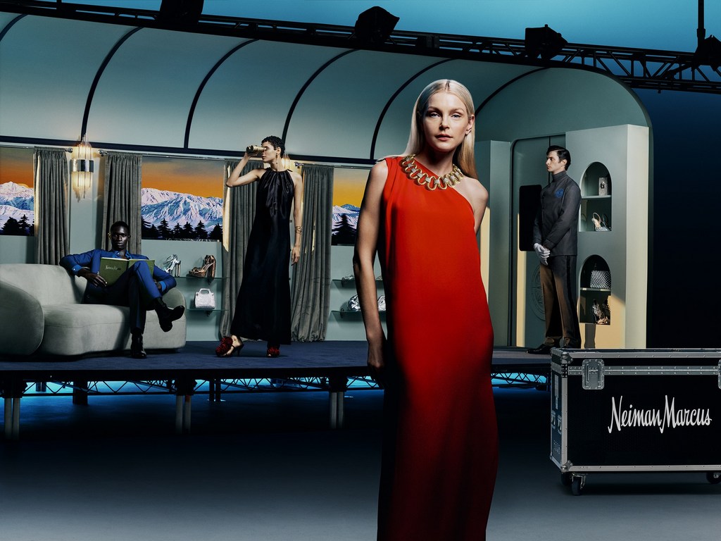 Fashion editorial shot in a staged setting: a woman in a red dress poses in front of a prop train carriage and luggage with the Neiman Marcus logo.