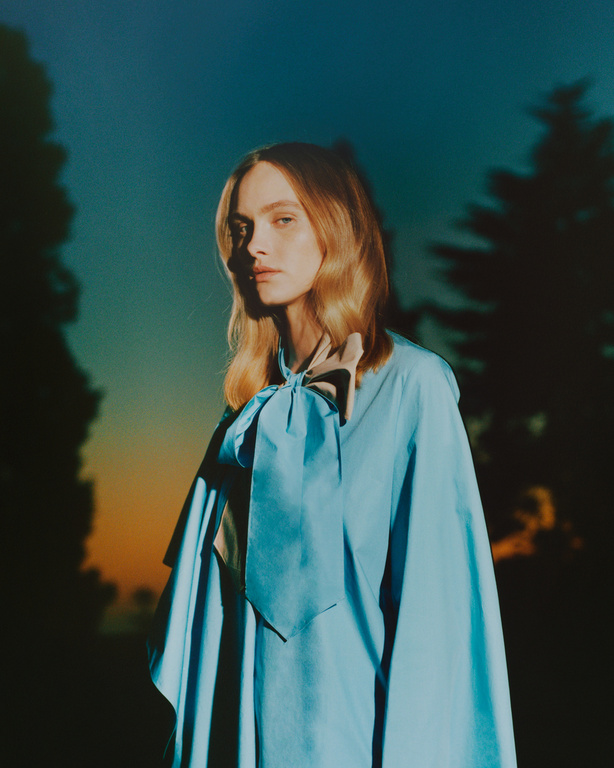 Model with blonde hair wearing a bright blue bow blouse against a dark twilight sky and tree silhouettes