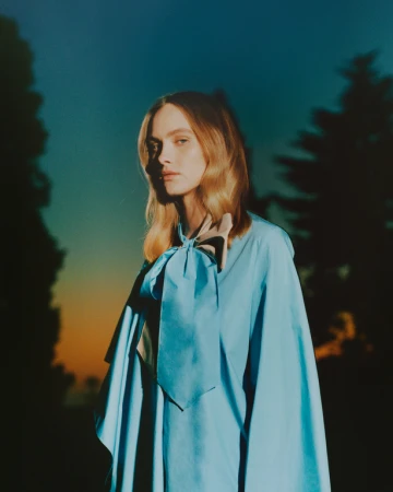 Model with blonde hair wearing a bright blue bow blouse against a dark twilight sky and tree silhouettes