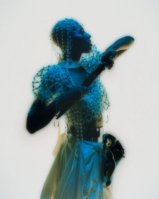 Person in beaded headpiece and shoulder piece, holding a dark object, in an artistic, studio-lit portrait against a white background.