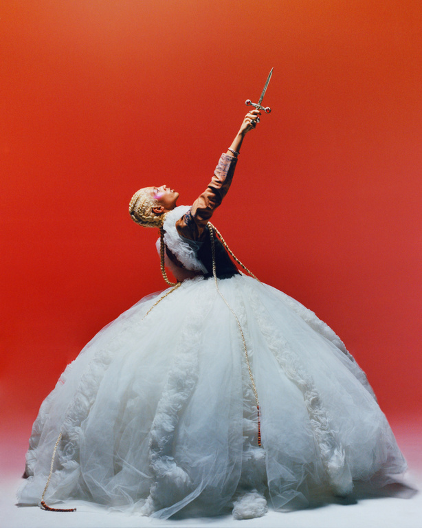 Woman in a large white gown holds a sword high against an orange background, creating a dramatic and visually striking image.