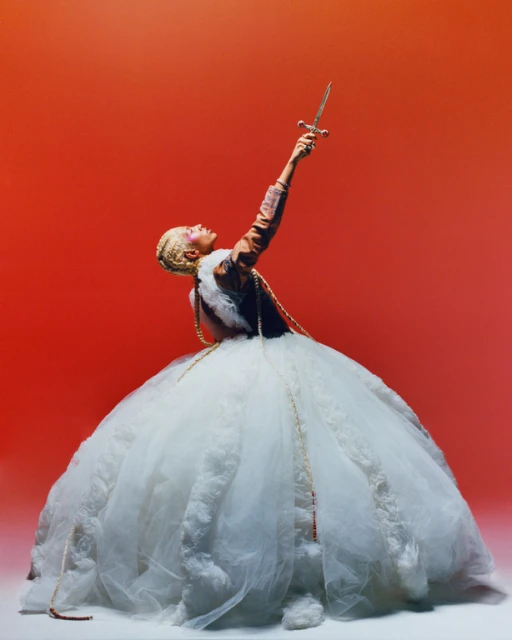 Woman in a large white gown holds a sword high against an orange background, creating a dramatic and visually striking image.