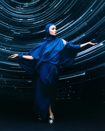 A woman in a striking blue gown and head covering poses against a dynamic, swirling starfield background, creating a fashion-forward image.
