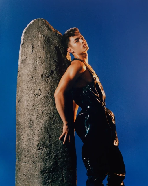 Stylish adult male model leans against a stone column in a studio with a blue backdrop, wearing a glossy black jumpsuit.