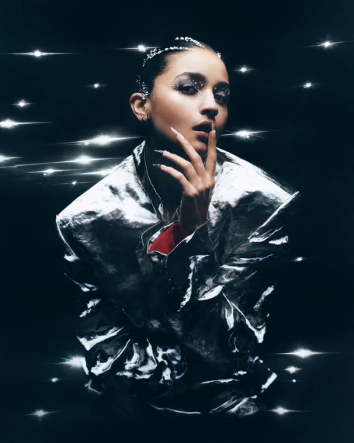 Glamorous portrait of a young adult female model with glitter makeup wearing a shiny silver jacket and posing in a dark studio.