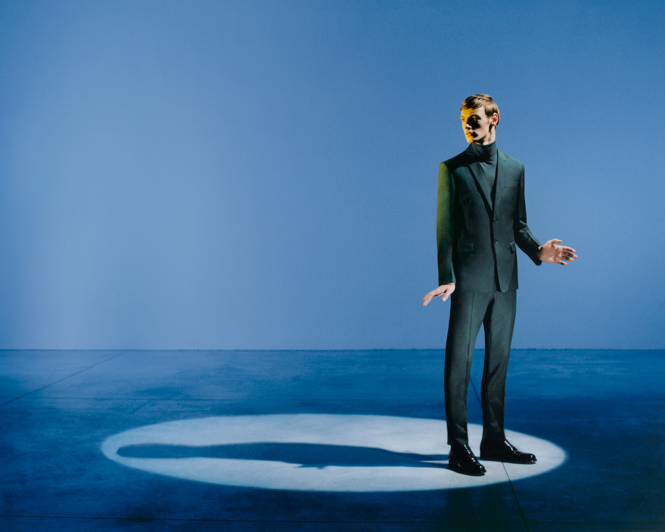 Man in dark suit stands in spotlight against solid blue backdrop, fashion portrait. Sharp focus, studio lighting, and modern aesthetic.