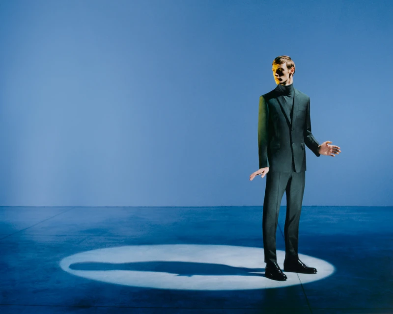 Man in dark suit stands in spotlight against solid blue backdrop, fashion portrait. Sharp focus, studio lighting, and modern aesthetic.