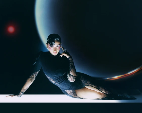 Model in black lace dress reclining against a surreal celestial backdrop with a glowing crescent planet and red light source