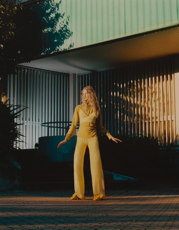 A slender woman with curly blonde hair poses outdoors in a mustard-colored, flowing pant suit with her arms outstretched in natural light.