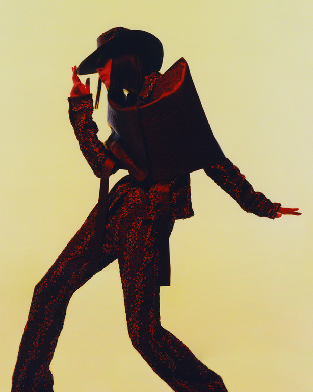 Fashionable person wearing a sequin suit and large hat strikes a pose in a studio setting, showcasing a modern and avant-garde style.