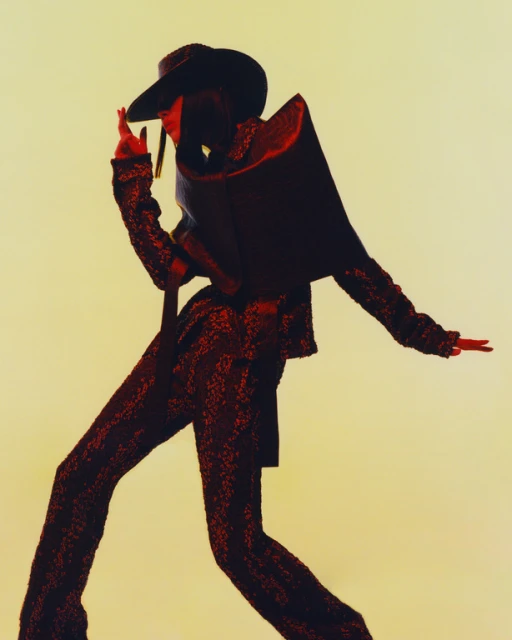 Fashionable person wearing a sequin suit and large hat strikes a pose in a studio setting, showcasing a modern and avant-garde style.
