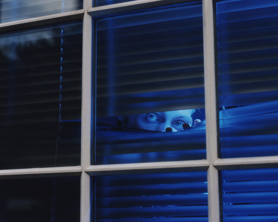Eye-level shot through blue blinds reveals a person's eyes peering out, illuminated in cool tones from an unseen light source.