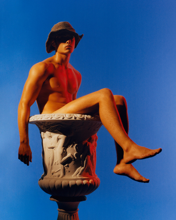 A nude man in a hat sits inside a stone vase against a blue background, lit with red light, highlighting musculature and form.