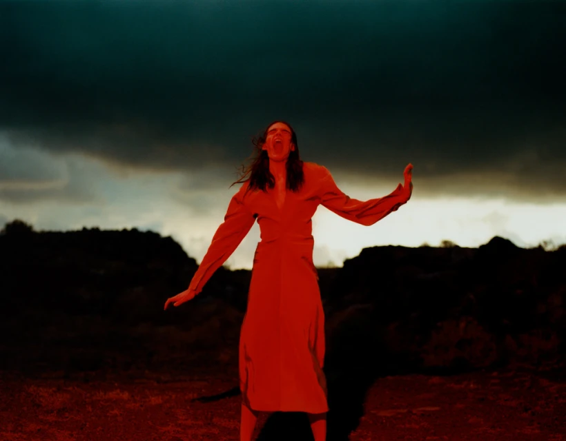 A woman in a red dress stands outdoors with her arms raised and face tilted upwards against a dark and stormy sky backdrop.