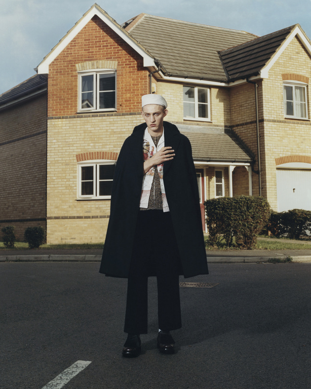 A stylish adult male stands on a street in front of a brick house, wearing a black coat, white sailor hat, and fashionable patterned shirt.