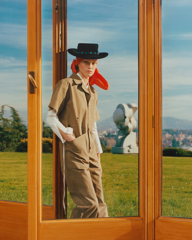 Fashion shot of a woman in a brown jumpsuit, black hat, and orange scarf, posed in a doorway overlooking a green lawn and cityscape.