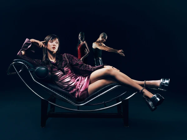 A woman in a purple dress and silver heels reclines on a black lounge chair in a dark studio with two figures blurred in the background.