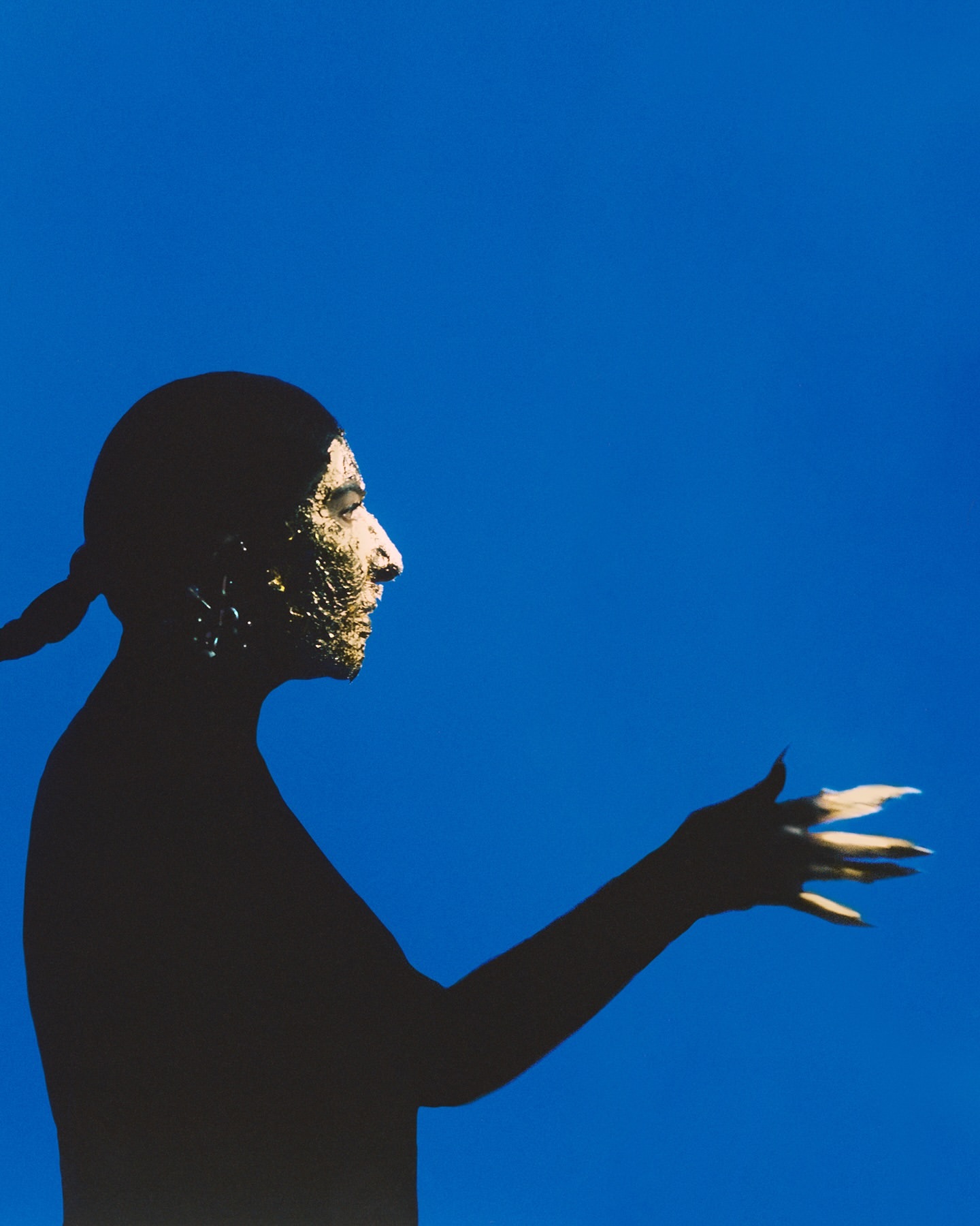 Side profile portrait of a person with textured skin and long nails against a saturated blue background, creating a striking contrast.