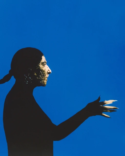 Side profile portrait of a person with textured skin and long nails against a saturated blue background, creating a striking contrast.