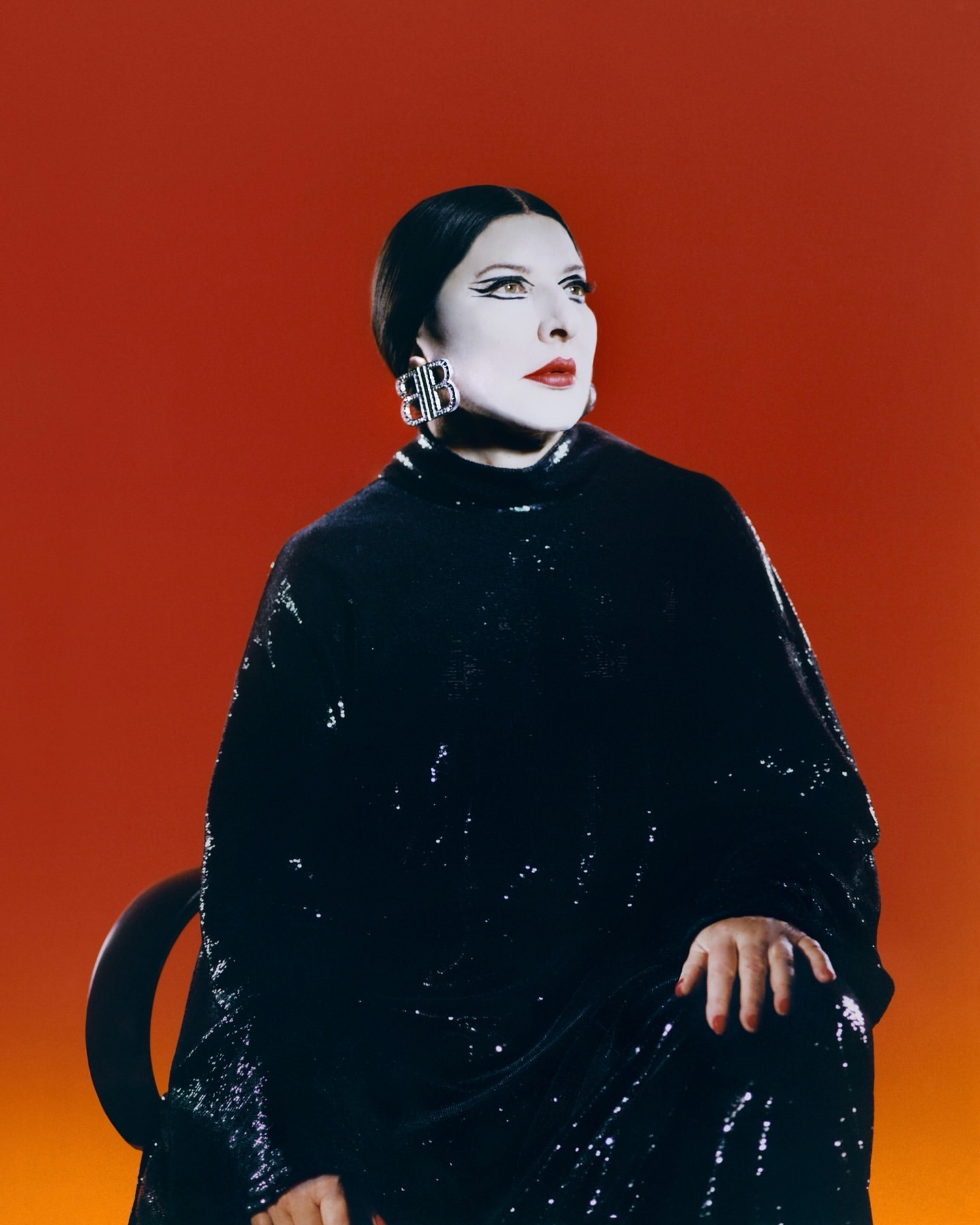 Elegant woman in a black sequin dress and statement earrings poses against an orange backdrop, looking up and away from the camera.