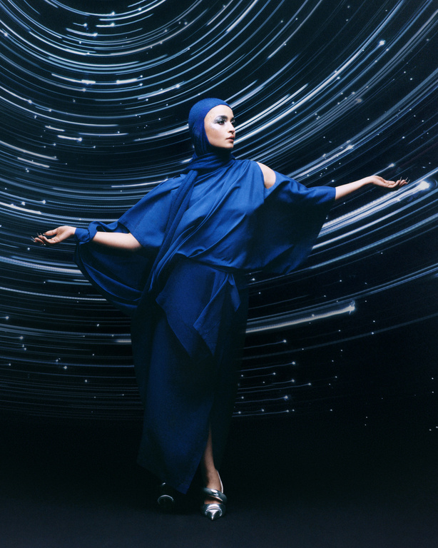 Elegant woman in a blue dress with head covering poses against a backdrop of blurred white light streaks in a studio setting.