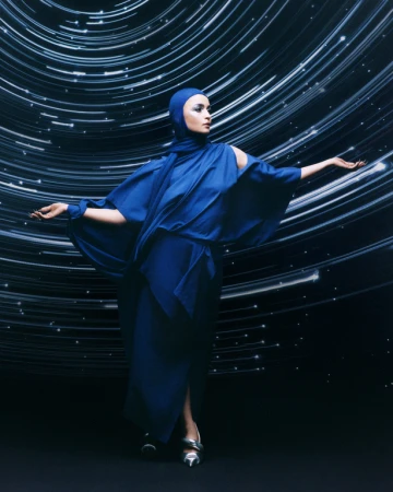 Elegant woman in a blue dress with head covering poses against a backdrop of blurred white light streaks in a studio setting.