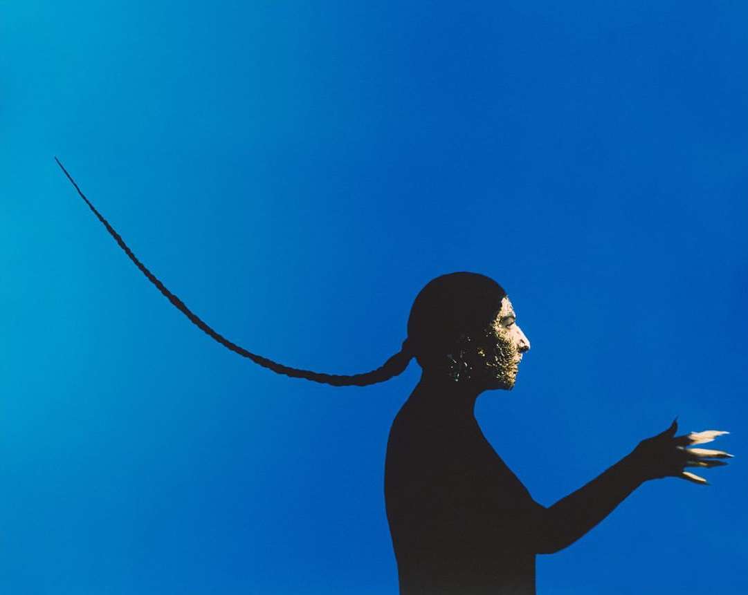 Artistic studio shot: Silhouetted person with ornate face paint and stylized braid against a vibrant blue background, distinctive visual.